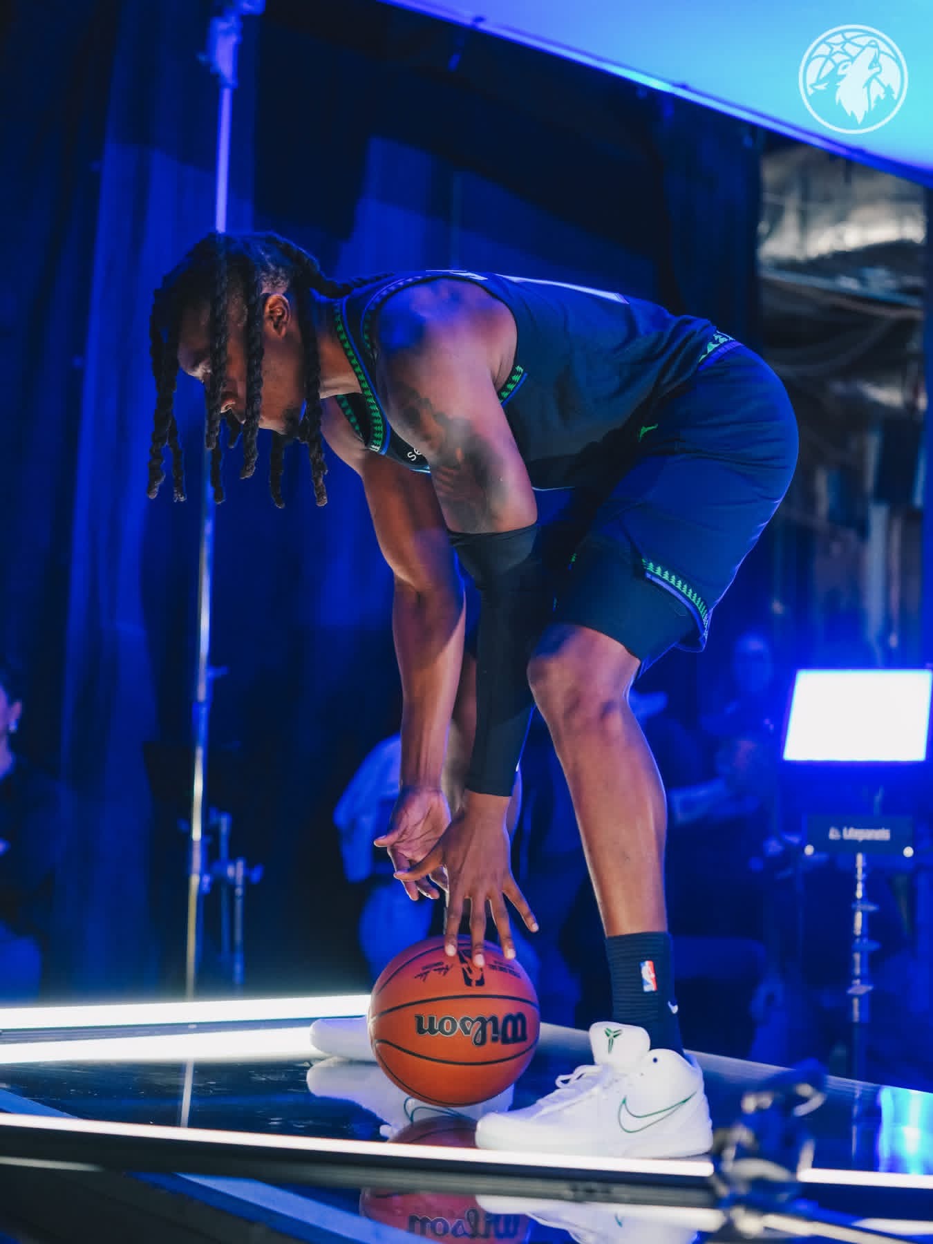 A Wolves player with long braids bending down to grab a basketball on a lit stage in the black city uniform